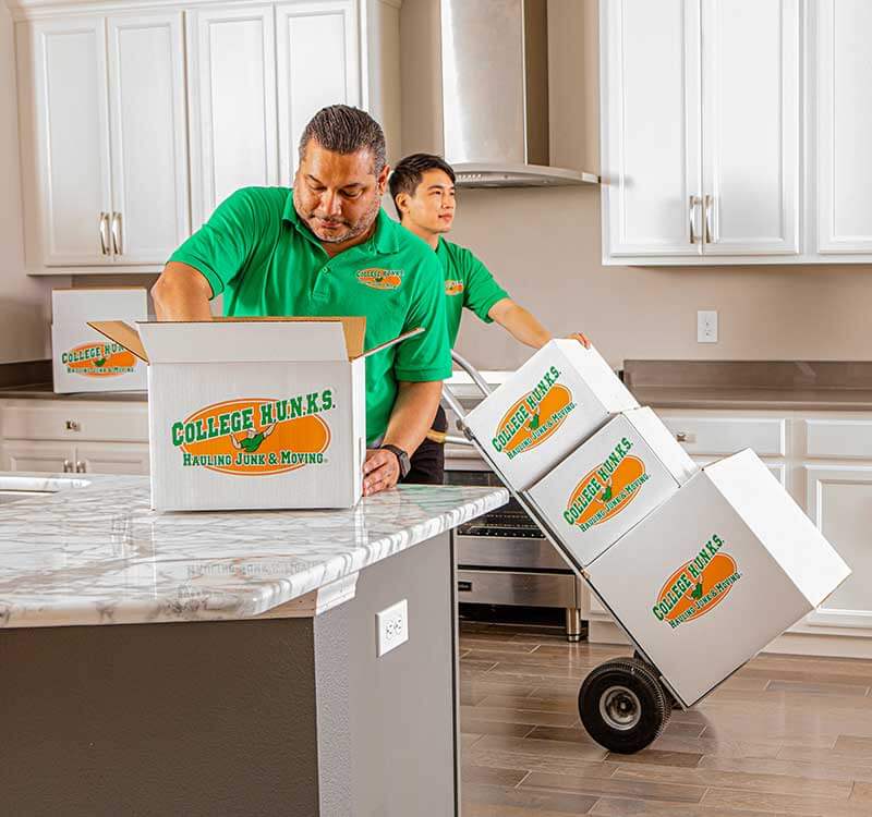 Two uniformed College HUNKS crew members packing labeled boxes inside a home