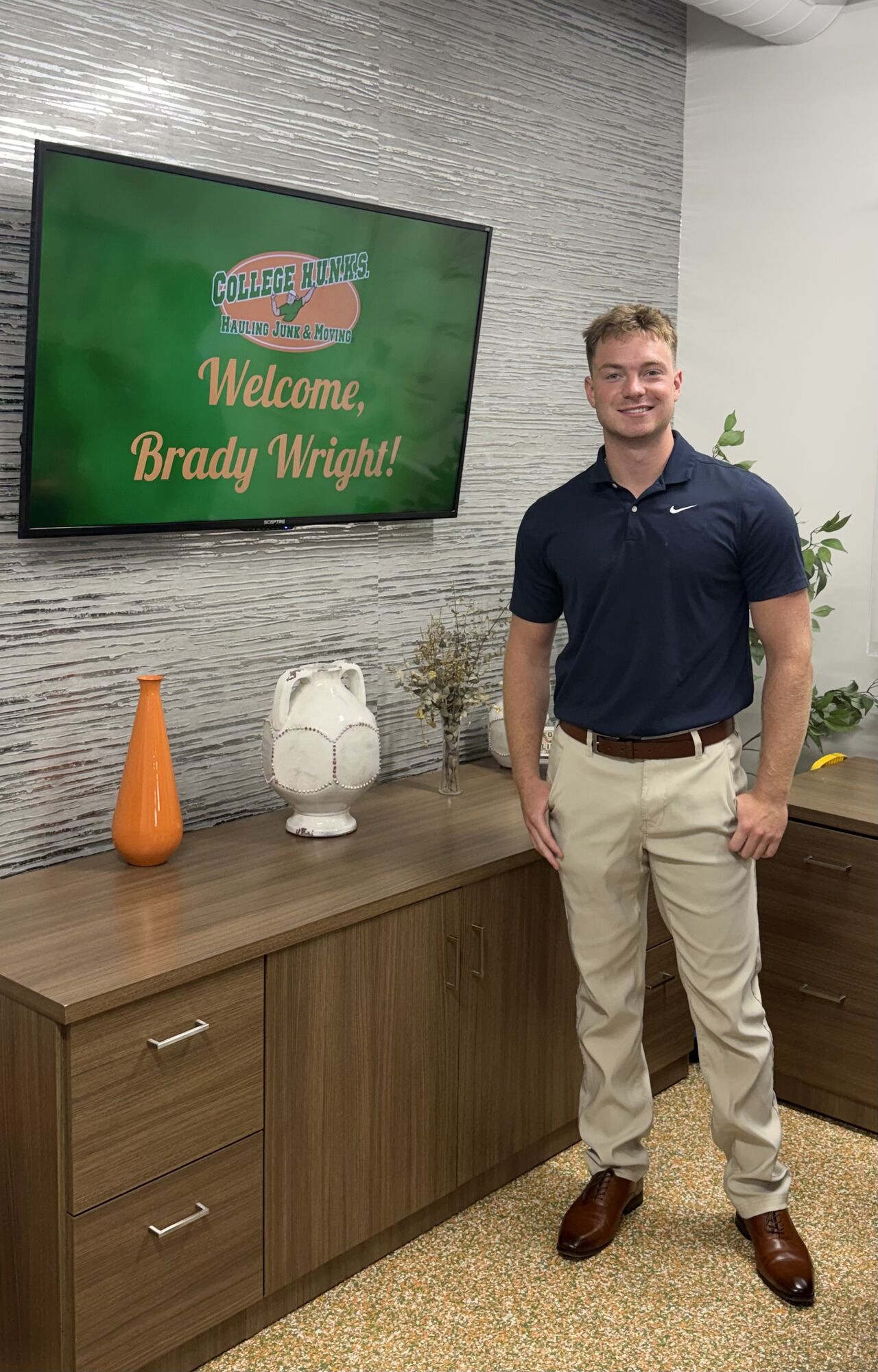 Brady Wright standing beside a Welcome, Brady Wright! screen at College HUNKS headquarters in Tampa, Florida